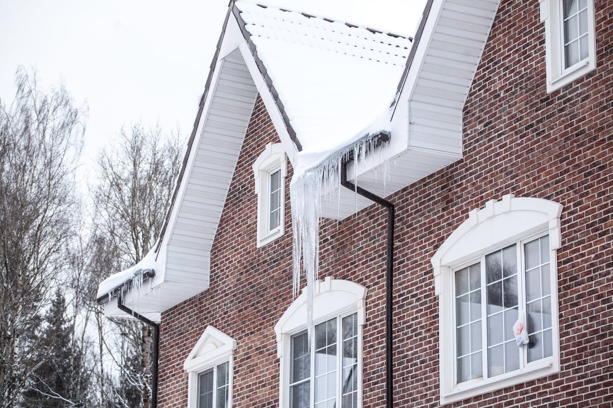 Close-up of a home's roof eave showing large icicles hanging and a thick ridge of ice built up against the shingles, with visible discoloration indicating moisture.