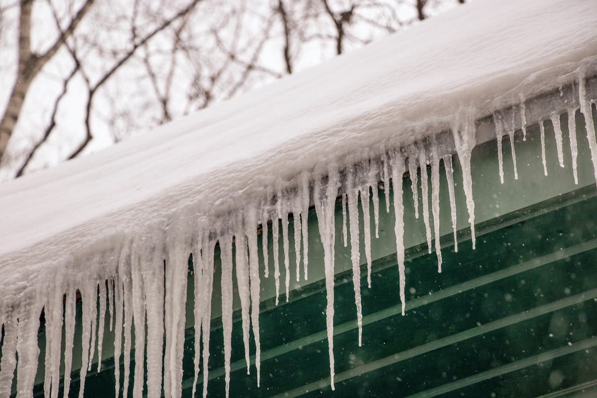 Cross-section diagram view of a roof showing snow accumulation at the ridge melting and refreezing at the eave to form an ice dam, with arrows indicating water intrusion path under shingles.